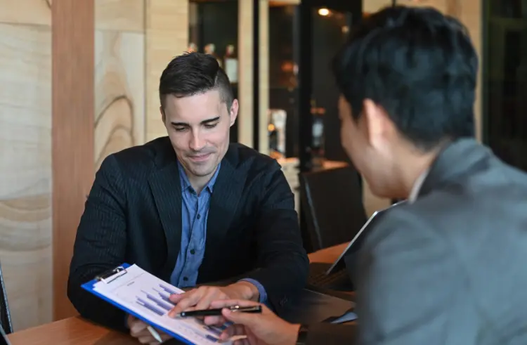 Two people in a business setting discuss a chart on a clipboard. One points to the document while the other listens. Both are sitting at a table with a laptop nearby.