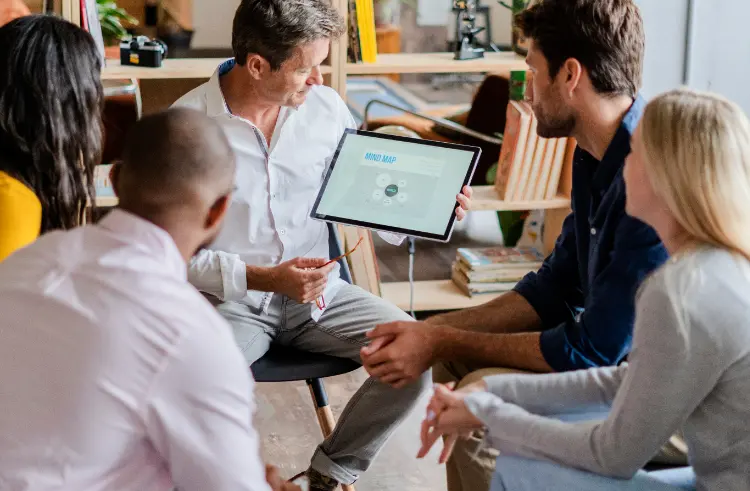 Five people have a discussion, with one person holding a tablet displaying a mind map. They are seated in a casual office setting.
