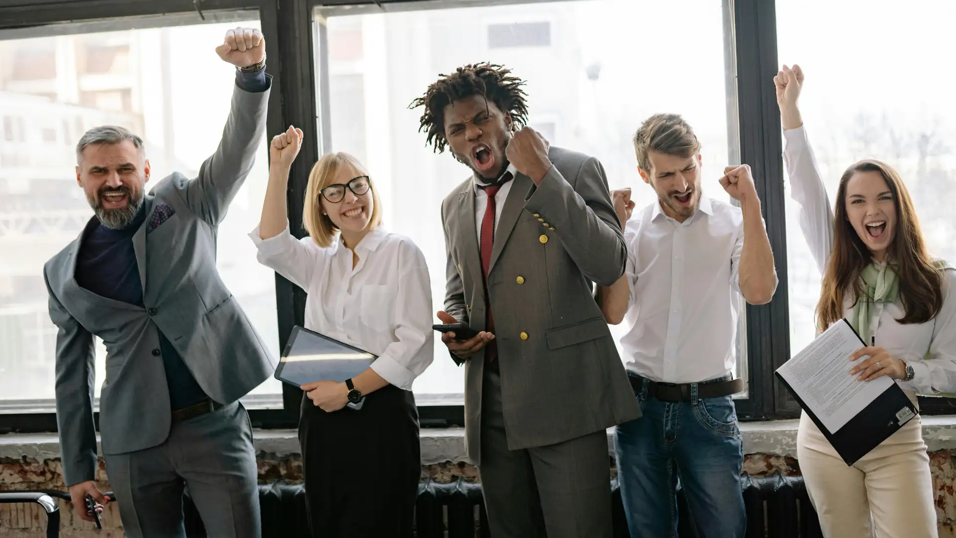 Five people in business attire stand indoors, smiling and raising their fists in celebration. With papers and a phone in hand, they embody the spirit of employee ownership, radiating success as they embrace their shared achievements.