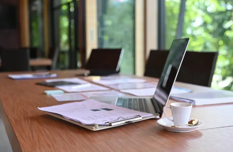 Open laptop on a wooden table with papers, documents, and a small cup of coffee. Sunlight streams through large windows in the background.