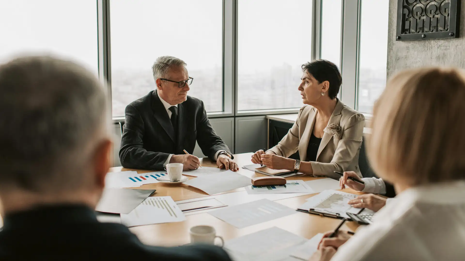 Business meeting with four people sitting around a conference table discussing documents, with a city view through large windows in the background.