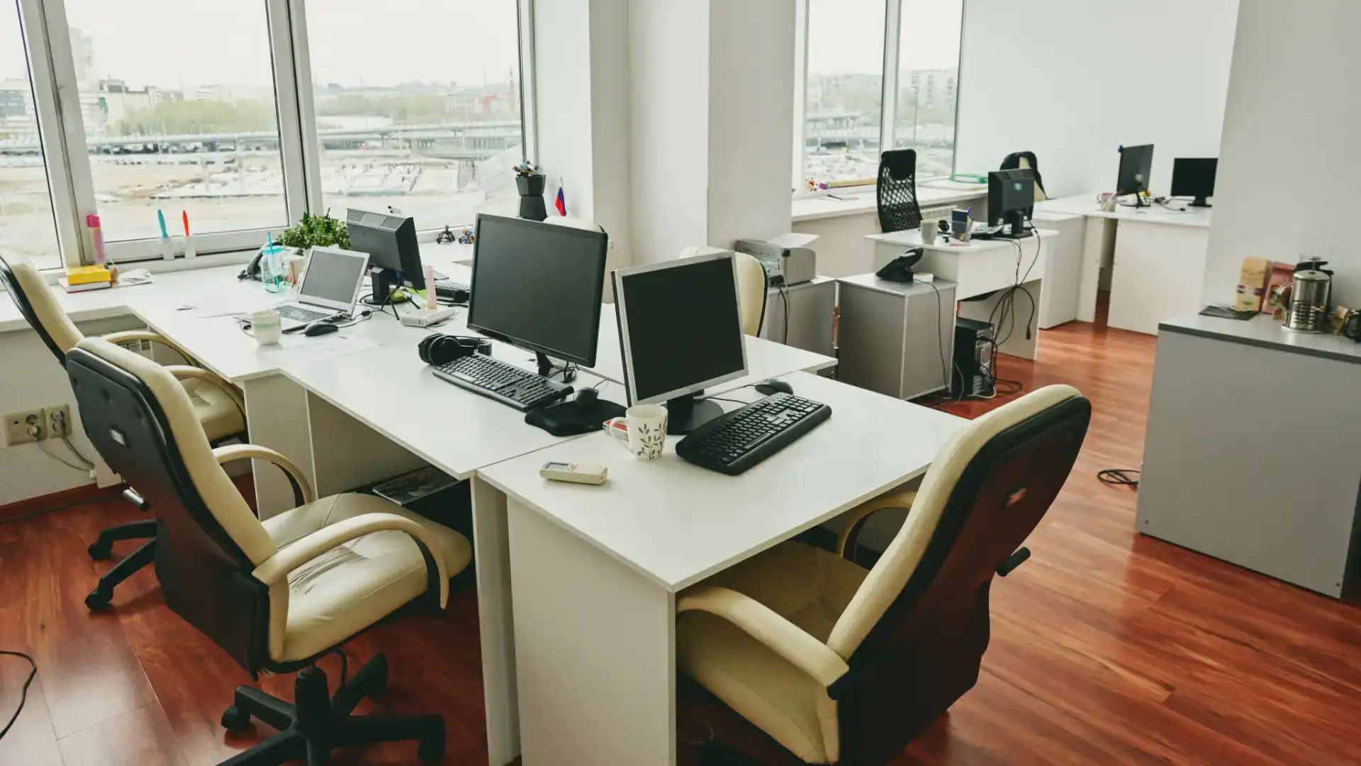 An empty office with multiple desks and chairs, each with a computer and keyboard. Large windows in the background provide natural light, and the floor is hardwood.