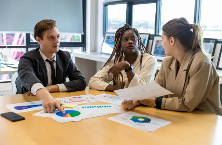 Three people sit at a table with charts and graphs, discussing work.