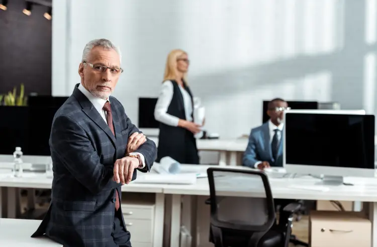 A man in a suit checking his watch stands in an office with two colleagues working at desks in the background.